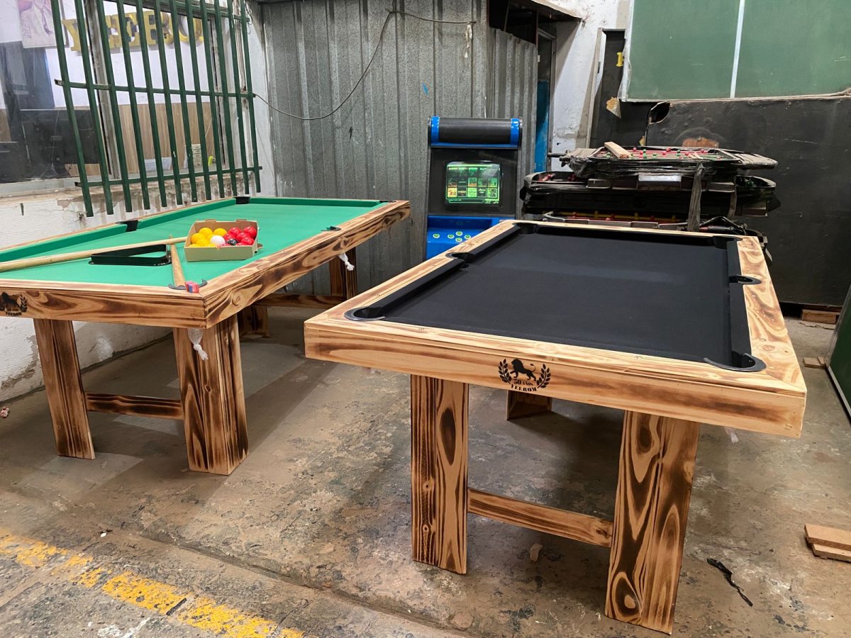 Dos mesas de billar de madera clara, una con paño verde y otra con paño negro, dentro del taller.